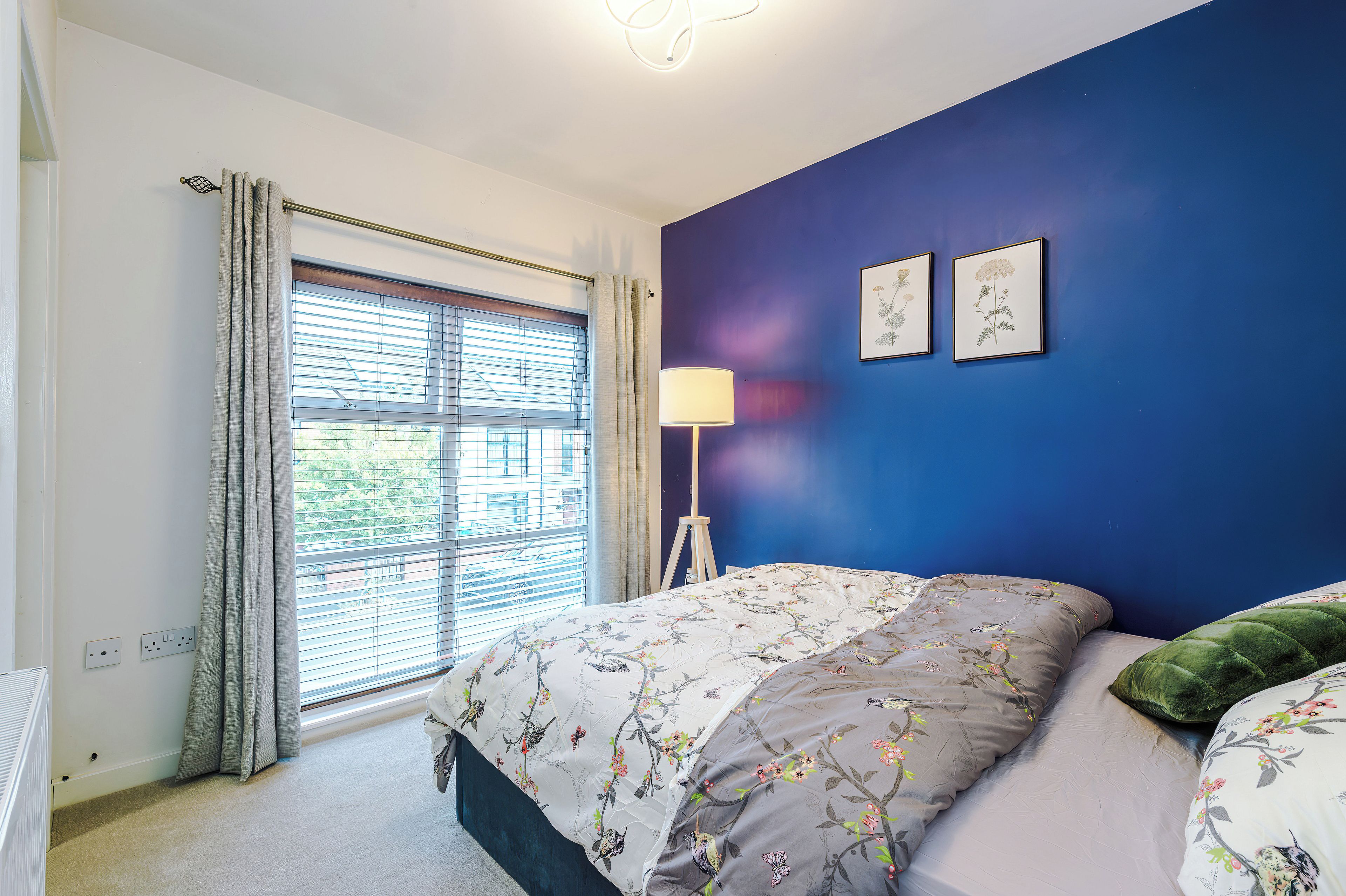 Une chambre à coucher avec un lit, une lampe sur pied et deux reproductions botaniques encadrées accrochées au mur.