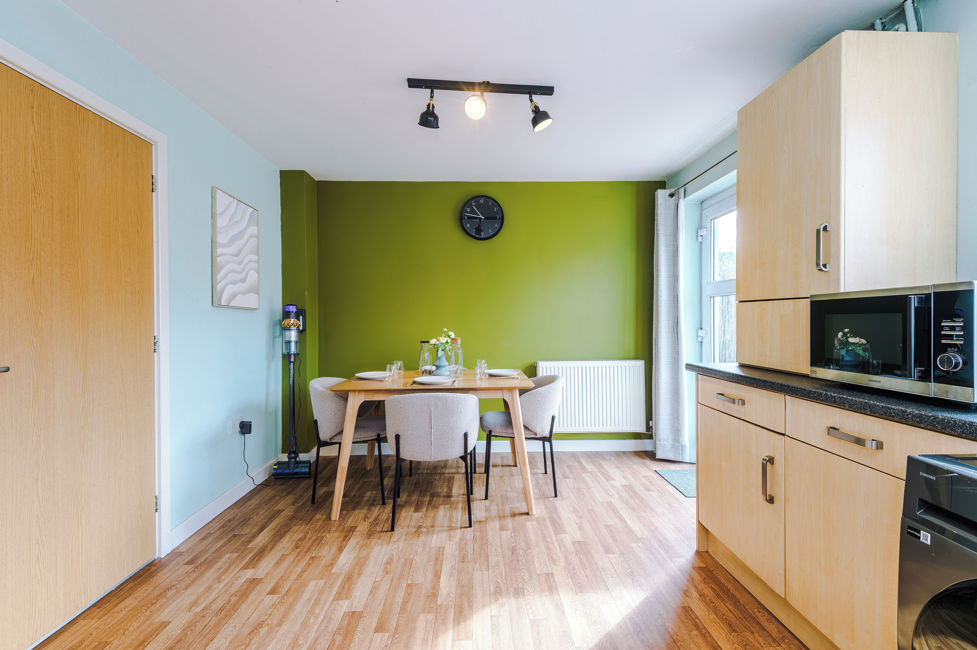 A compact kitchen with a dining table, chairs, a clock, and a microwave.