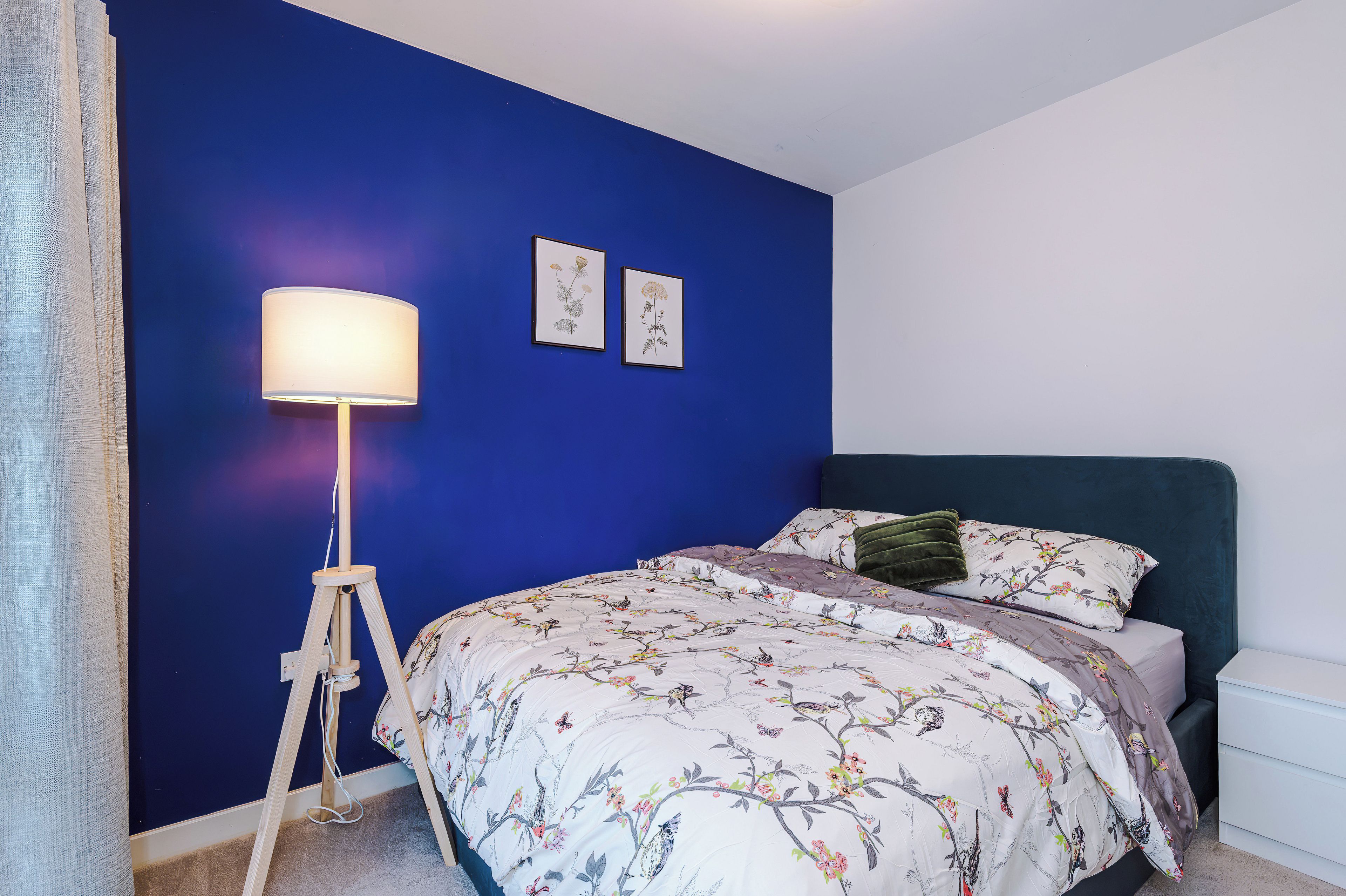 Une chambre avec un mur bleu, un lit recouvert d’une couette à motifs floraux, une lampe sur pied et deux illustrations botaniques encadrées.