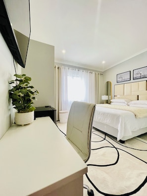 A hotel room with a bed, a desk, a chair, a television mounted on the wall, and a potted plant.