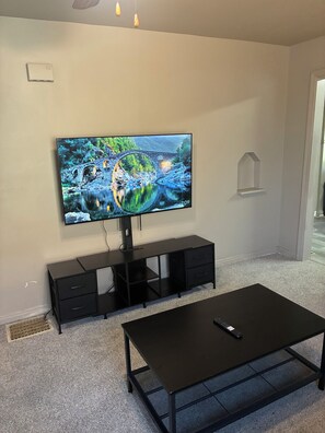 Ein Wohnzimmer mit einem an der Wand befestigten Flachbildfernseher, einer schwarzen Unterhaltungsanlage darunter und einem schwarzen Couchtisch im Vordergrund.
