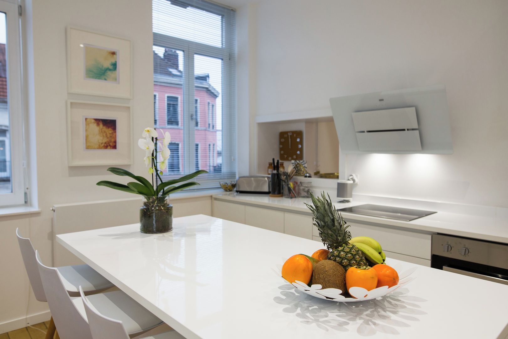 Une cuisine moderne avec des plans de travail blancs, un panier de fruits, une orchidée en pot et une fenêtre donnant sur la ville.