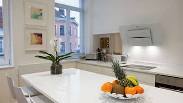 Une cuisine moderne avec des plans de travail blancs, un panier de fruits, une orchidée en pot et une fenêtre donnant sur la ville.