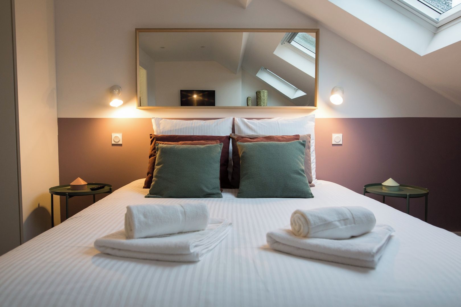 Une chambre d’hôtel avec un grand lit, deux tables de chevet, un miroir et une fenêtre de toit.