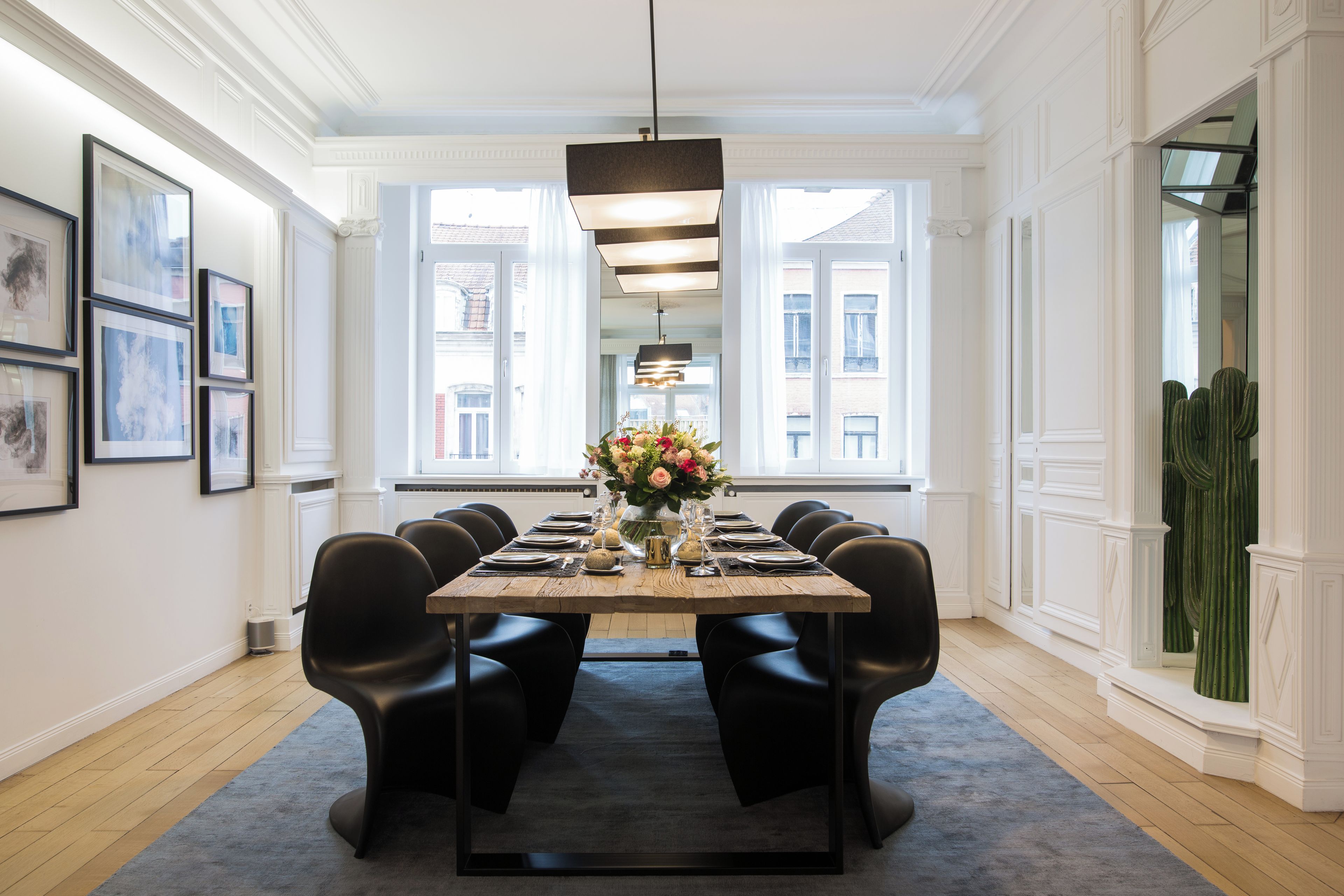 Une salle à manger avec une longue table dressée pour six personnes, entourée de chaises noires, un lustre au-dessus et une fenêtre donnant sur des immeubles.
