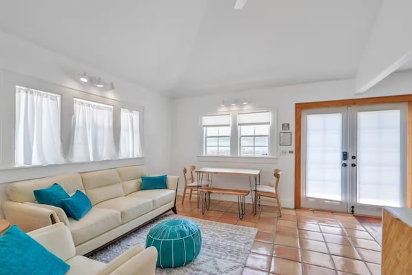 En rummelig opholdsstue med sofa, spisebord og stole. Store vinduer med gardiner, flisegulv og en dør med greb.