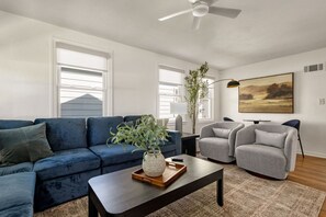 A modern living room with a blue sofa, grey armchairs, a coffee table, and a large window with blinds. - 3BR/1BA Updated South Bend Retreat Close to ND (South Bend)