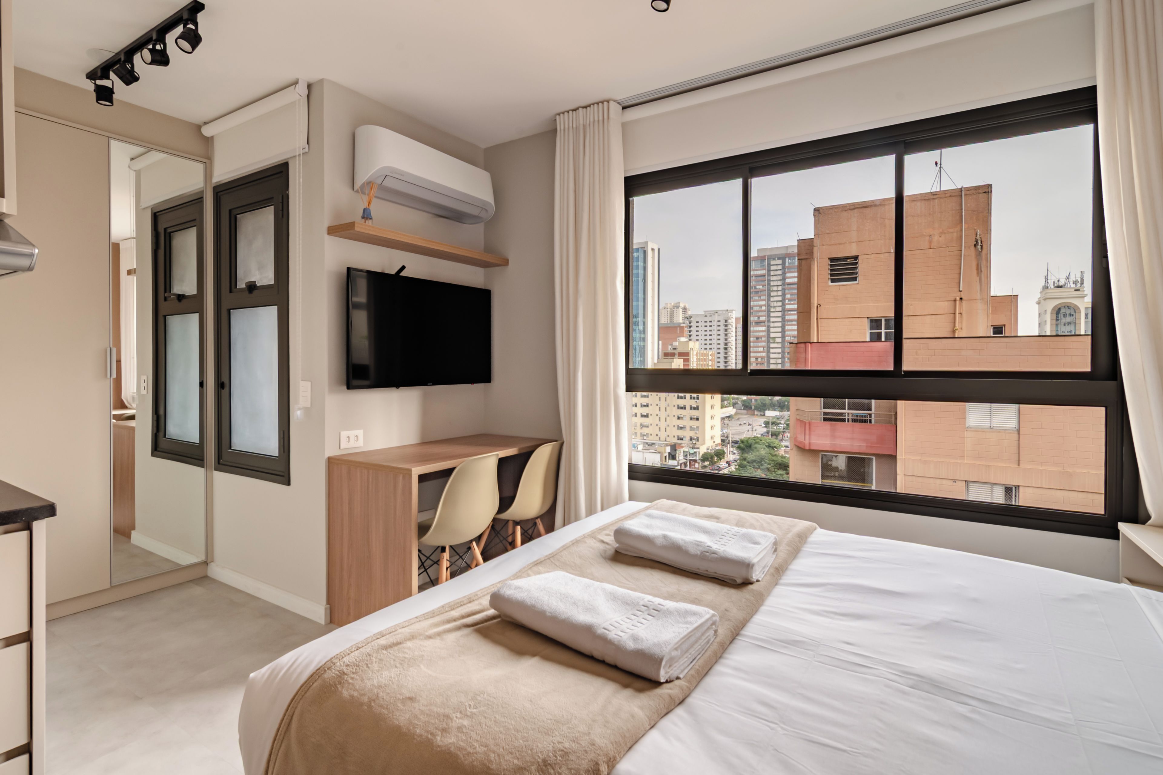 A hotel room with a bed, a desk, a chair, a TV, and a window with a city view.