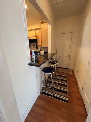 Een kleine keuken met een aanrecht, twee barkrukken, een houten kast en een deur.