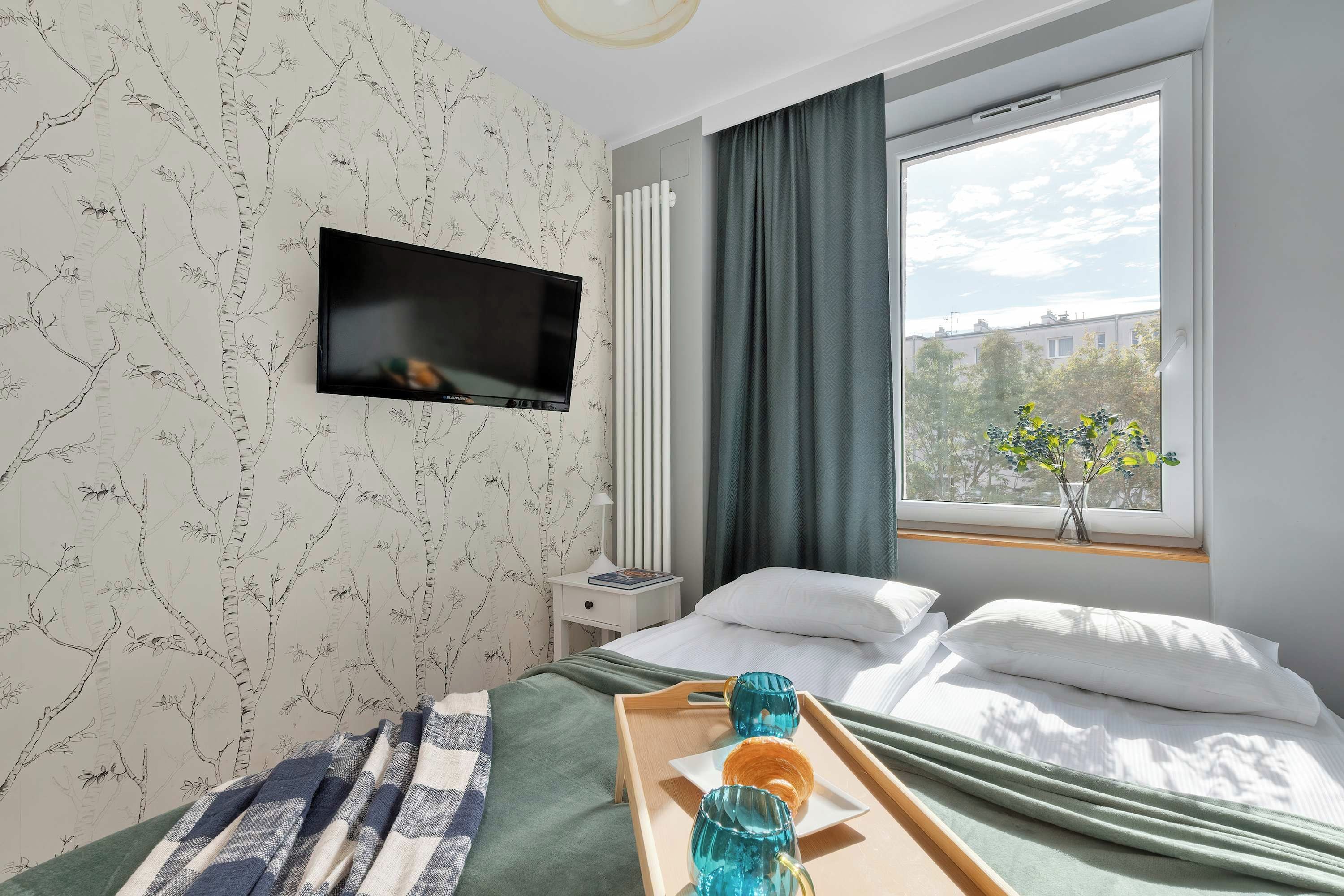 A hotel room with a bed, a TV mounted on the wall, a window with curtains, and a tray with a glass and a book.