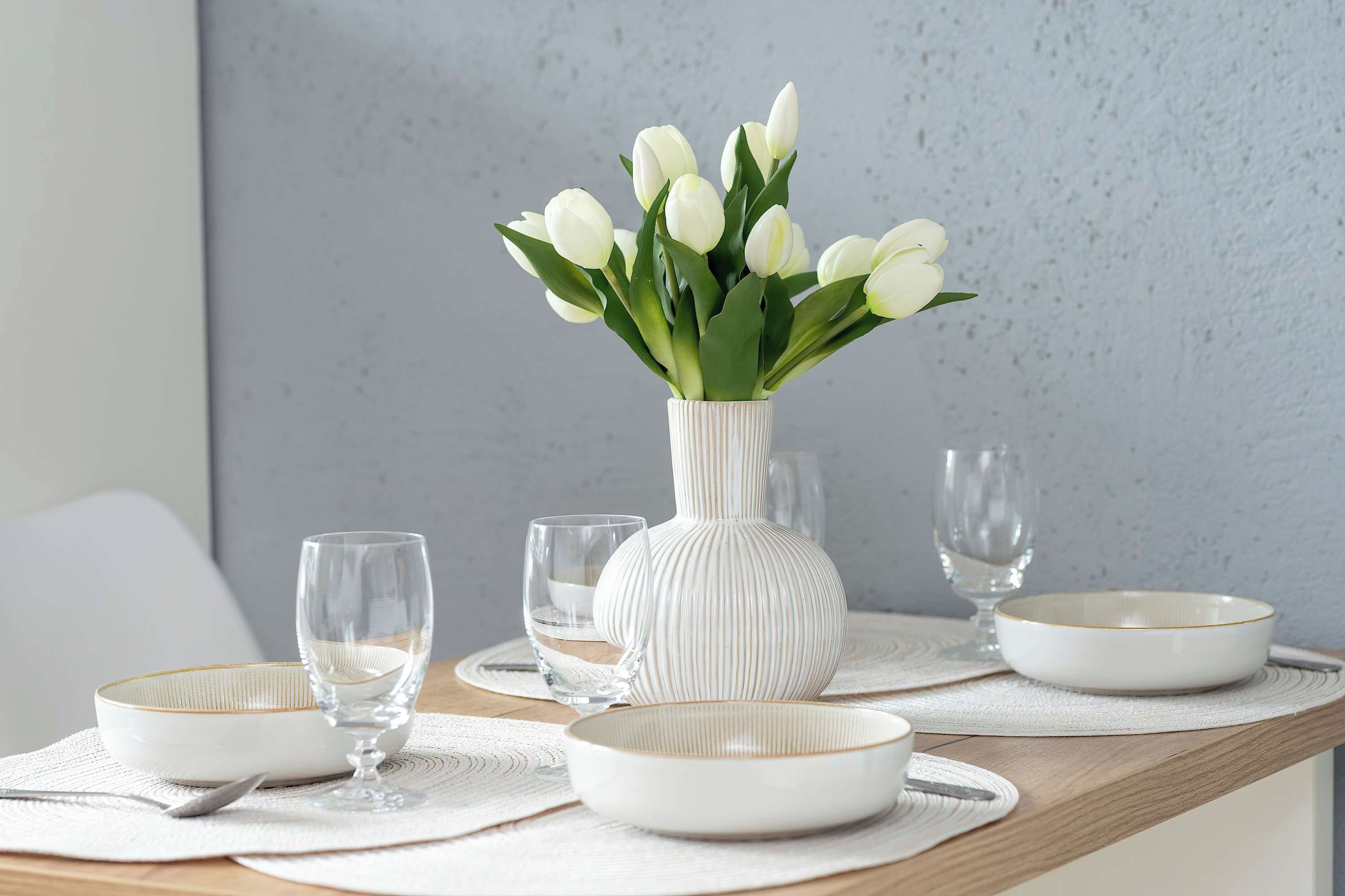 A dining table with white plates, bowls, and glasses, a white vase with white flowers, and a textured wall in the background.