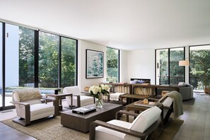 A spacious living room with large windows, a coffee table, sofas, and a bookshelf. - Shore House - A Luxury Beach Retreat (Shelter Island Heights)