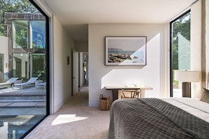 A modern bedroom with a large bed, a desk, and a view of a balcony with lounge chairs. - Shore House - A Luxury Beach Retreat (Shelter Island Heights)