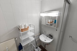 A bathroom with a white sink, a mirror, a shelf with toiletries, and a shower area. - A studio apartment just a 5-minute walk from the cable cars (Crans-Montana)