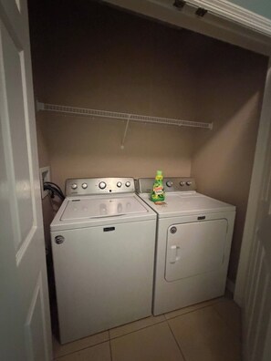 A laundry room with two white front-loading washers and a dryer, a bottle of Gain Softener, and a white shelf above the dryers. - Apartment*Disney*Universal in Windsor Palms (Kissimmee)