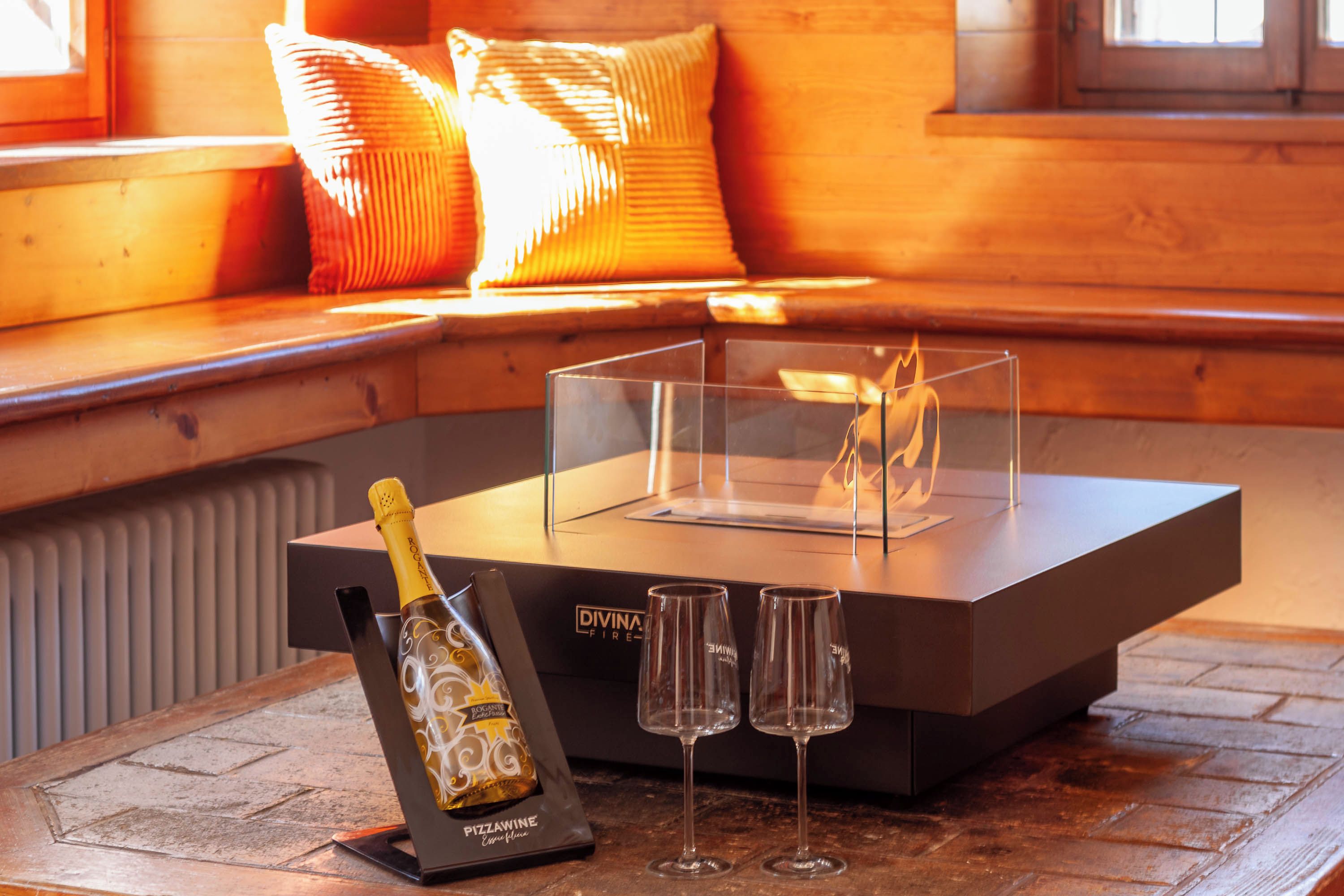 Un coin salon accueillant avec un banc en bois et des coussins, une table en verre transparent sur laquelle se trouvent une bouteille et des verres, et une cheminée.