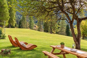 A picnic setup with a wooden table and two wooden chairs on a grassy hill with trees in the background.