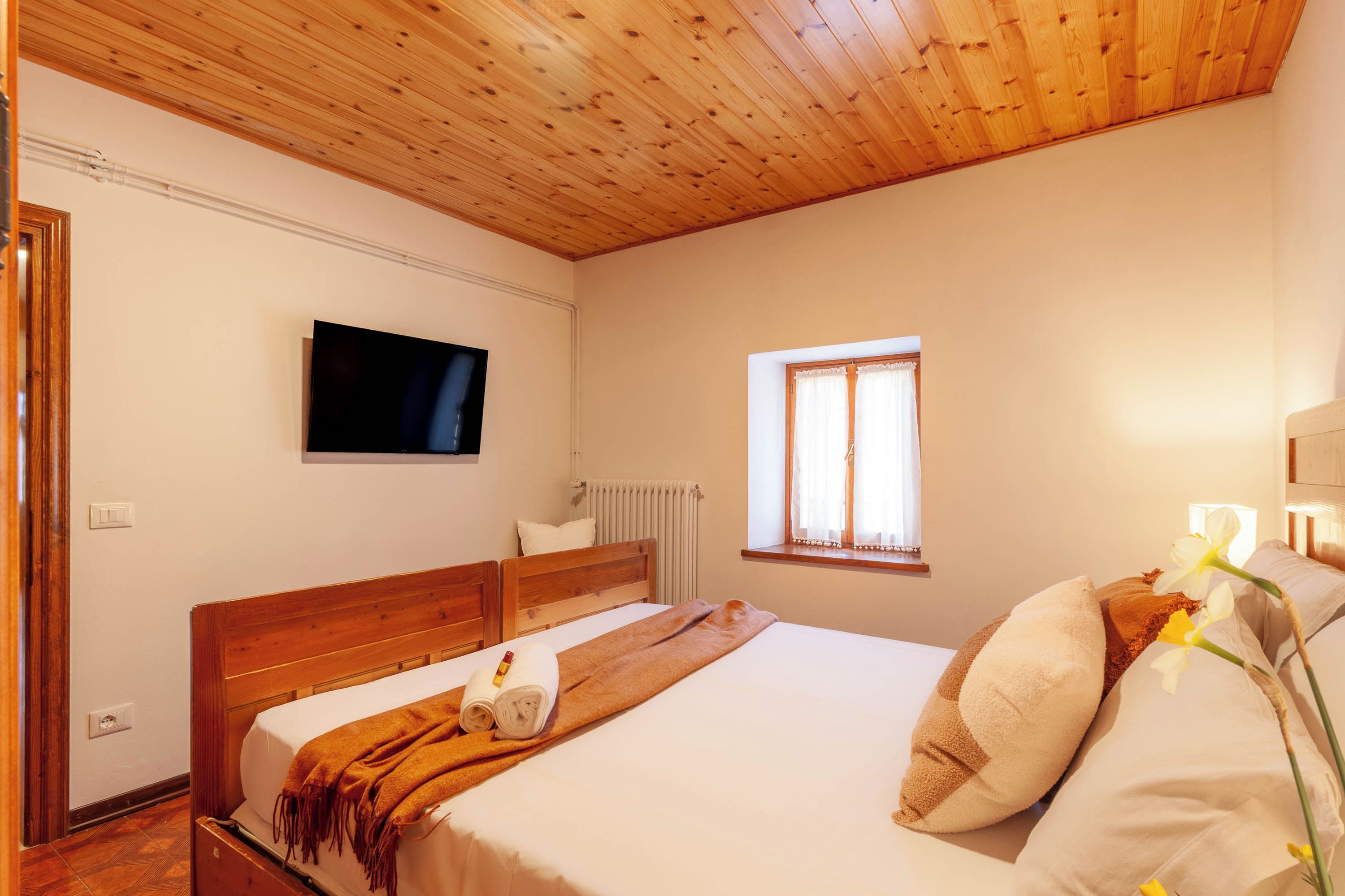 Une chambre avec un plafond en bois, un lit recouvert de draps blancs, un téléviseur fixé au mur et une fenêtre avec des rideaux.