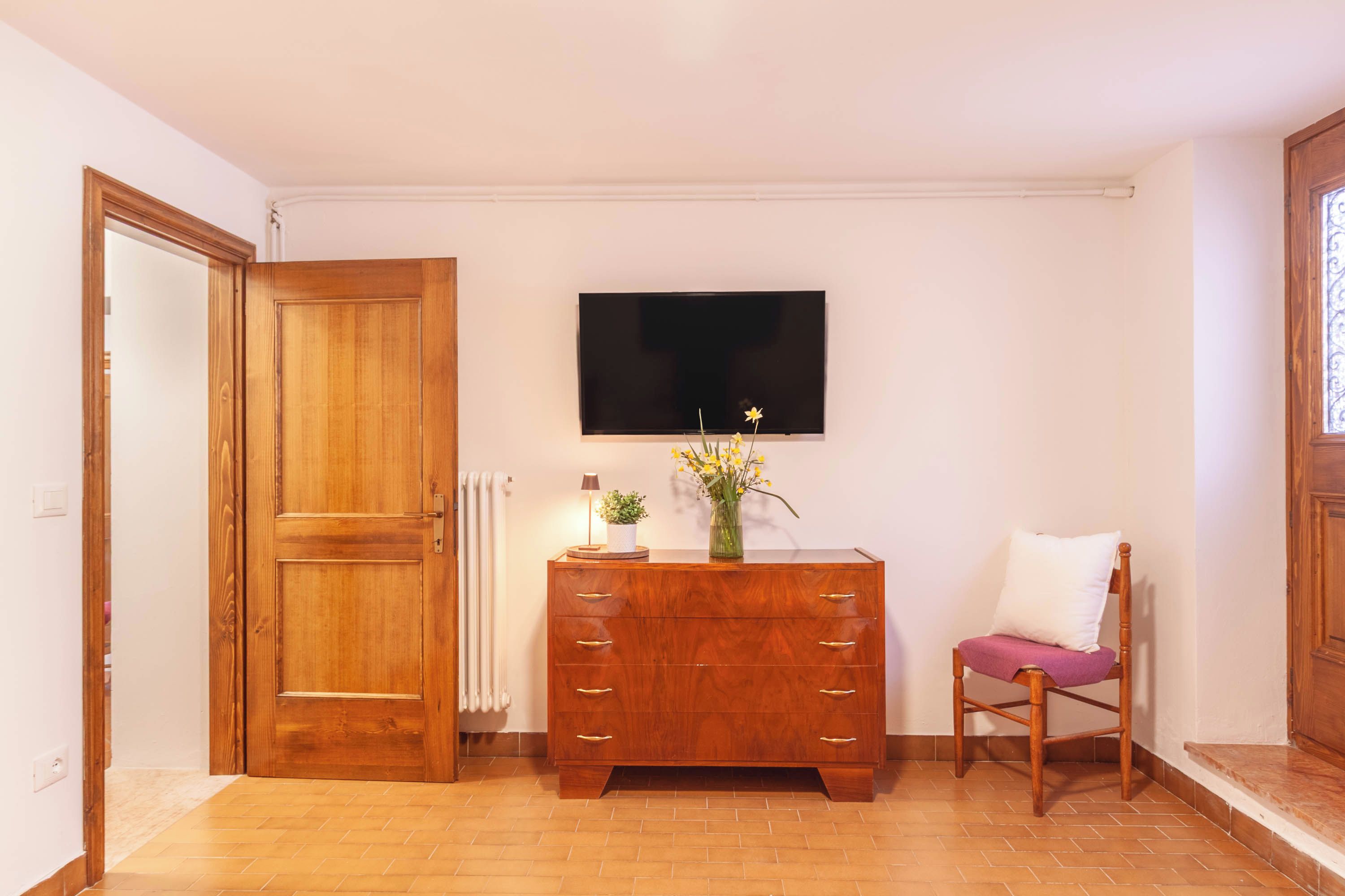 A room with a wooden door, a dresser with drawers, a chair with a cushion, a flat-screen TV mounted on the wall, and a radiator.
