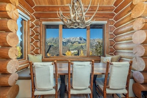 A log cabin interior with a dining area featuring a table and chairs, a large window with a mountain view, and a chandelier. - Newly Furnished 3BR Ski Townhome Sleeps 8 + Private Hot Tub (Mountain Village)