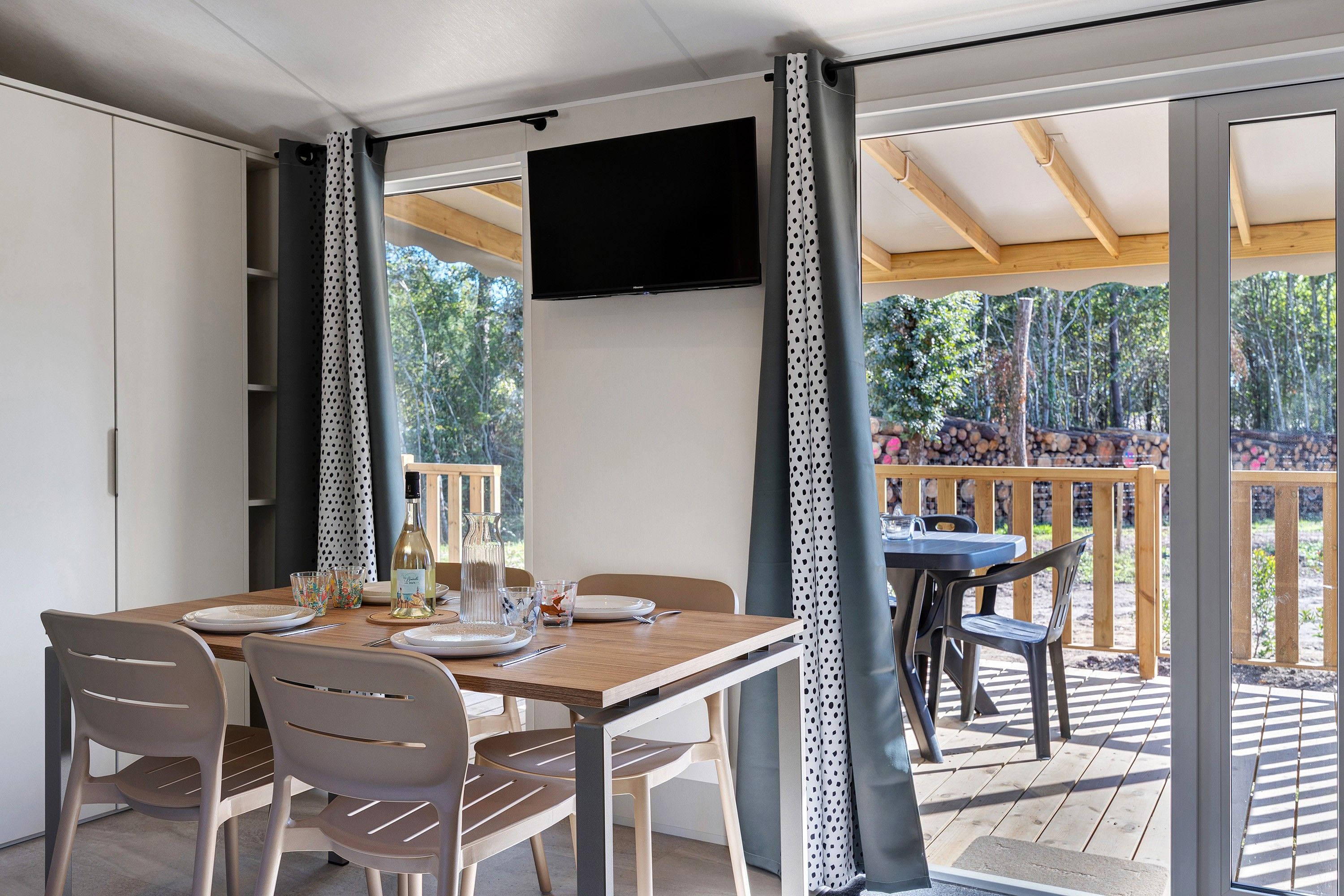 Ein Essbereich mit einem für vier Personen gedeckten Tisch, Stühlen, einem an der Wand befestigten Fernseher und Blick ins Freie durch große Fenster.