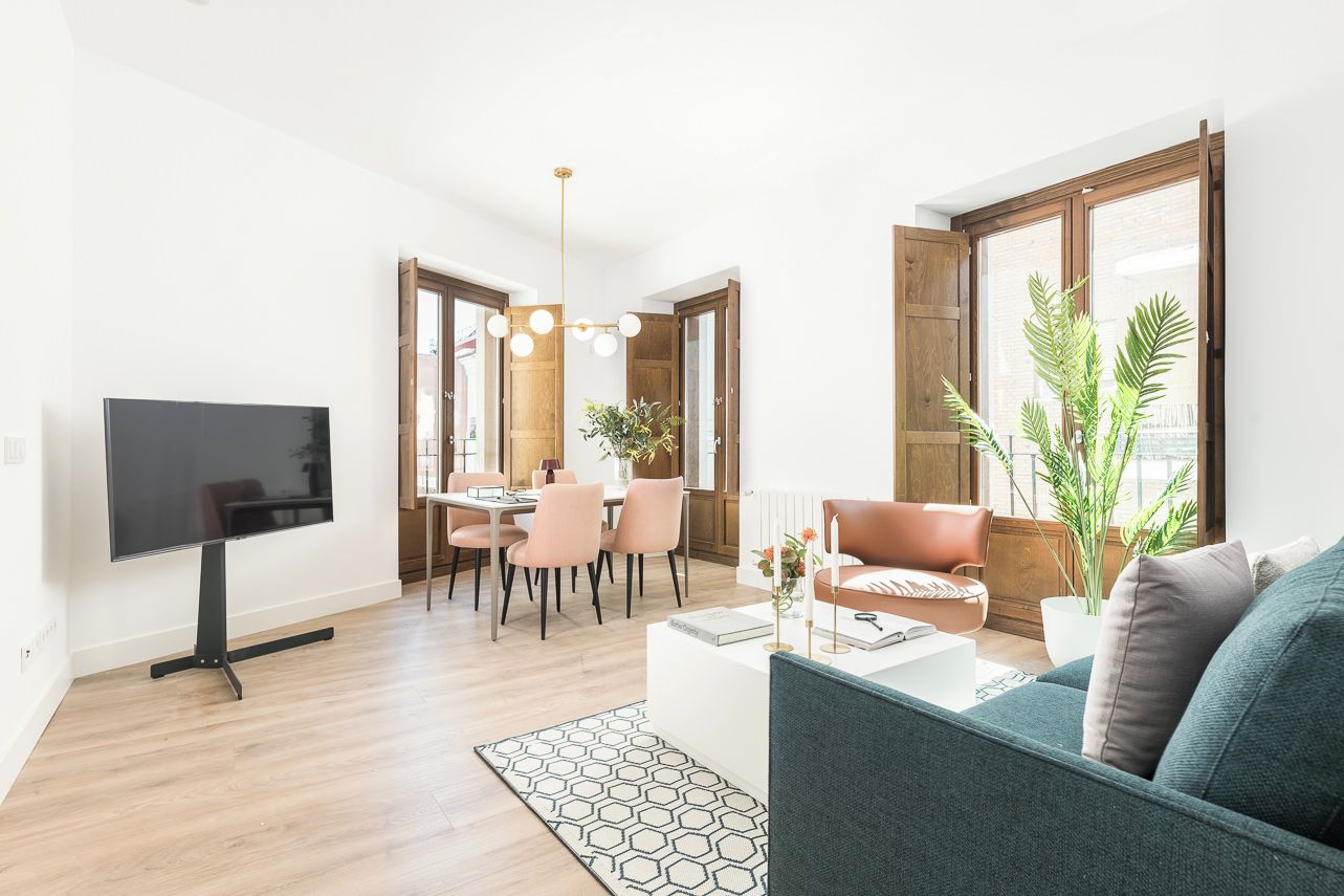 A modern living room with a flat-screen TV, a sofa, a dining area with chairs, and a large window with plants.