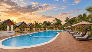 Zona de piscina en un resort con terraza de madera, sillas de descanso y palmeras.