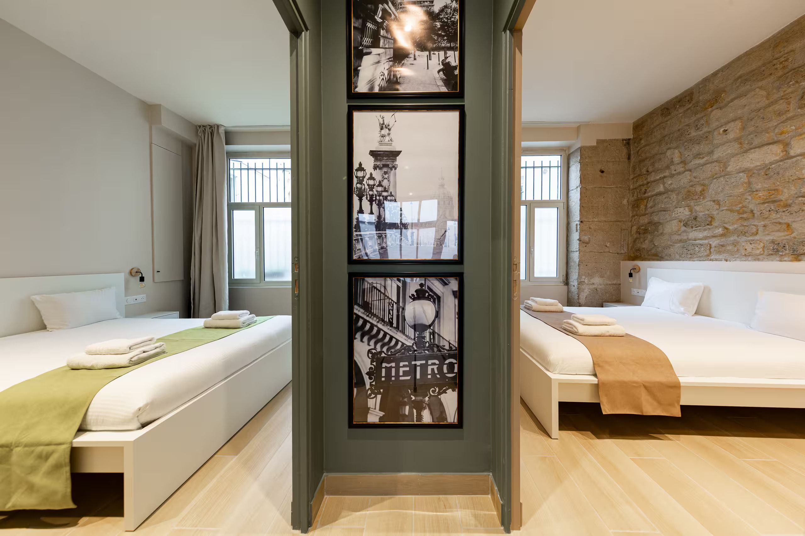 Une chambre d’hôtel avec deux lits, une colonne centrale ornée de tableaux encadrés, un mur en pierre et une fenêtre avec des rideaux.