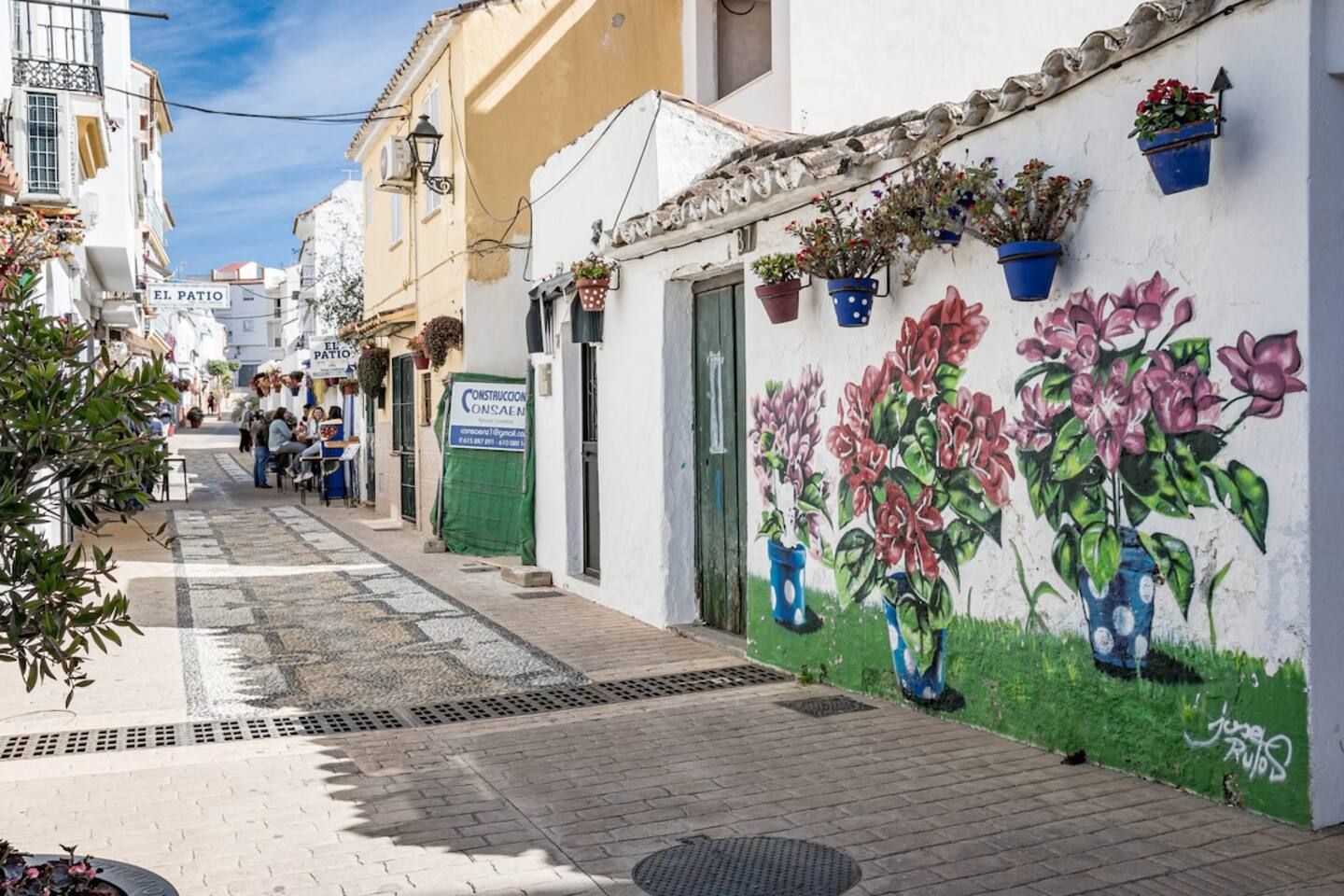 Een smalle straat met gebouwen aan weerszijden, een muurschildering van bloemen aan de rechtermuur, en een bordje met 'El Patio' aan de linkerkant.