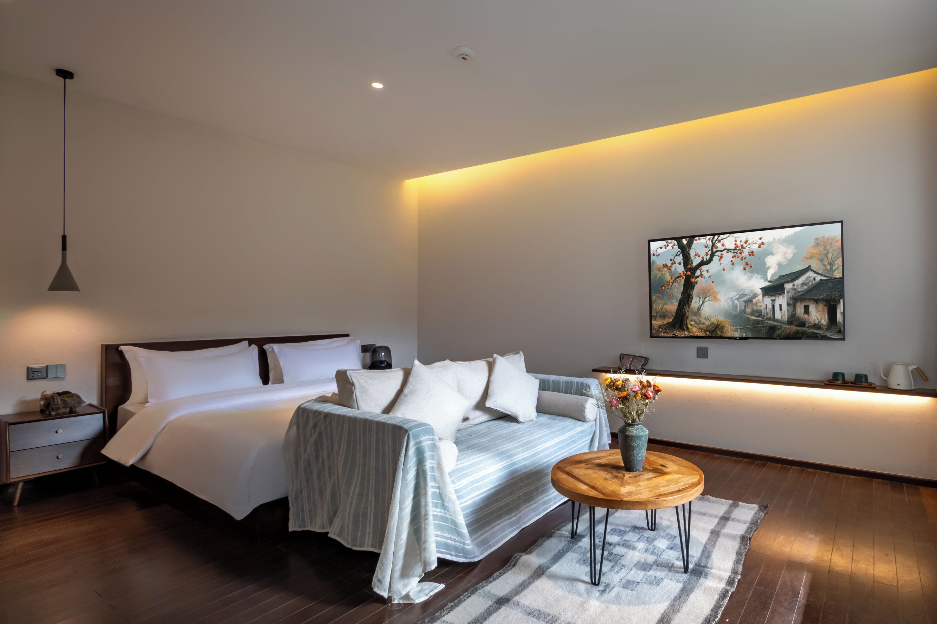 Een moderne slaapkamer met een bed, nachtkastjes, een lampje naast het bed, een klein rond tafeltje met een vaas, en een schilderij aan de muur.
