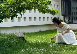 Una persona asseguda a l'herba prop d'una paret blanca amb una finestra.