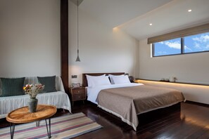 Quarto de hotel moderno com uma cama grande, um sofá, uma mesa pequena com um vaso de flores e uma janela com vista para o céu.