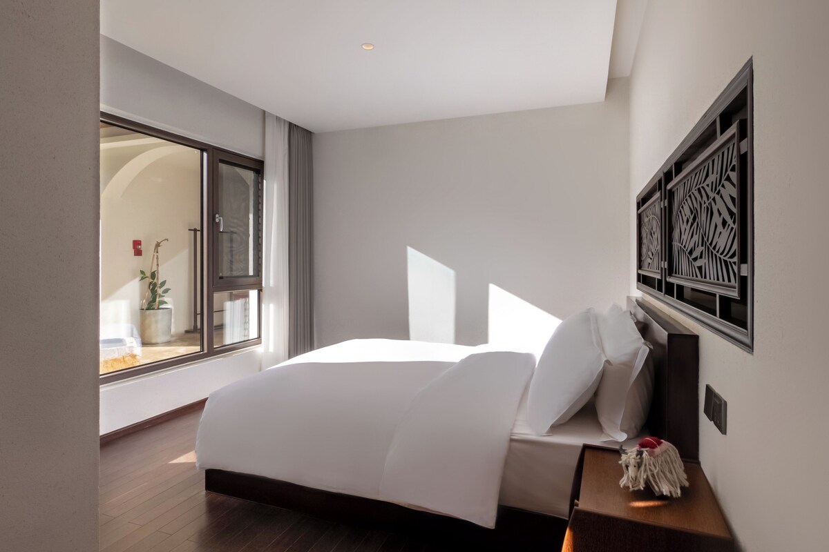 A hotel room with a large bed, a nightstand, a wall-mounted decorative panel, and a view through a window.
