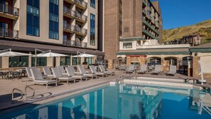 Ein Hotelpoolbereich mit Liegestühlen und Sonnenschirmen, ein klares, blaues Schwimmbecken und Blick auf die umliegenden Gebäude und Hügel.