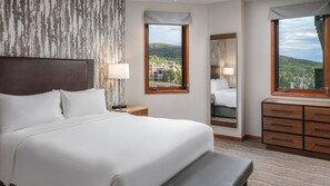 Ein Hotelzimmer mit einem großen Bett, Nachttischen, einem Schminktisch und einem Fenster mit Blick auf eine Berglandschaft.
