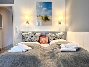 A hotel room with a bed, pillows, a headboard, wall-mounted lamps, and a painting of a beach scene. - Fiete Deppe House - Apt. 1 (Wyk auf Foehr)