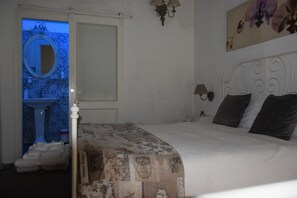 Un dormitori amb un llit, una tauleta de nit, una làmpada, un mirall fixat a la paret i un bany visible a través d'una porta oberta.