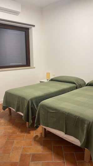 A room with two single beds, a window with blinds, a bedside lamp, and a tiled floor. - Green Home Apartment (Bolsena)