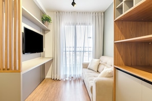A modern living room with a flat-screen TV mounted on the wall, a sofa with cushions, a wooden floor, and a large window with sheer curtains. - Becker by Bhomy (São Paulo)