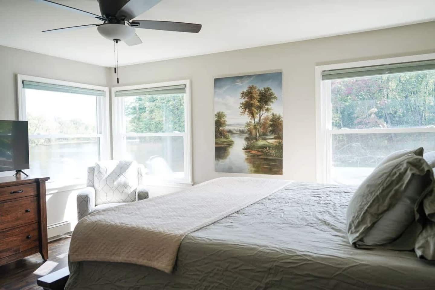 A bedroom with a bed, a ceiling fan, a dresser, a TV, and large windows with a view of trees and a lake.