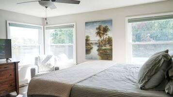 A bedroom with a bed, a ceiling fan, a dresser, a TV, and large windows with a view of trees and a lake.