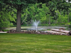 A tranquil park setting with a pond, a fountain, a picnic table, and a large tree.