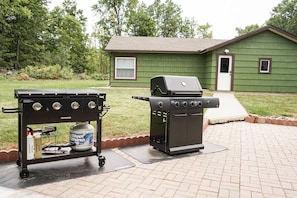 A barbecue grill and a gas tank on a paved patio with a green house in the background.