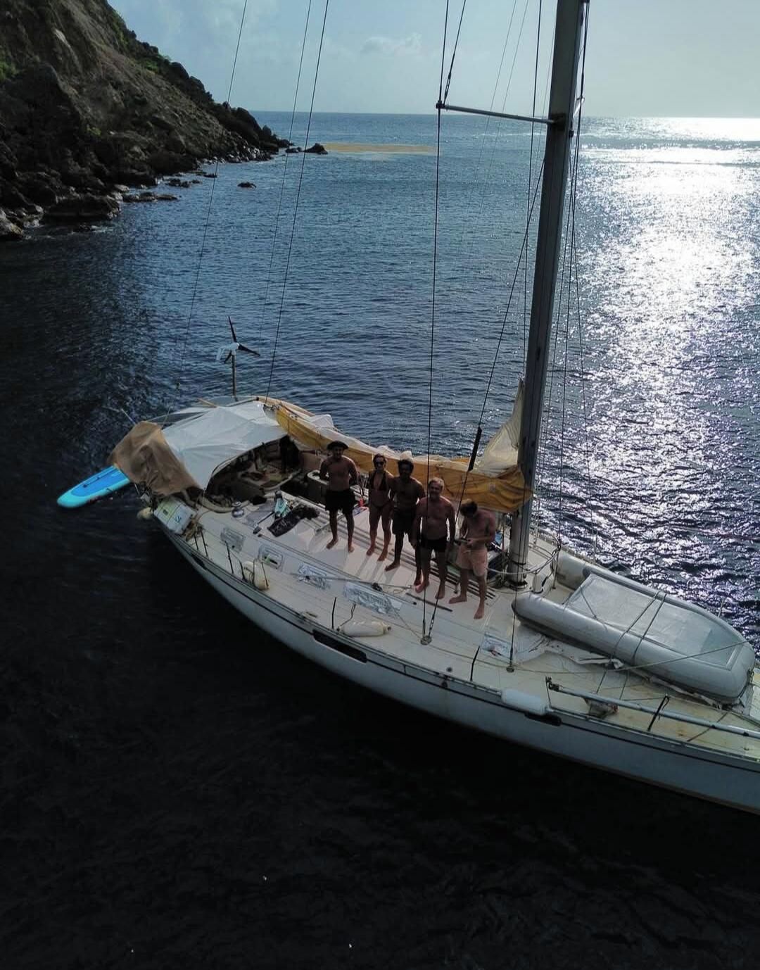 Un velero con un grupo de personas en la cubierta, anclado cerca de una costa rocosa.