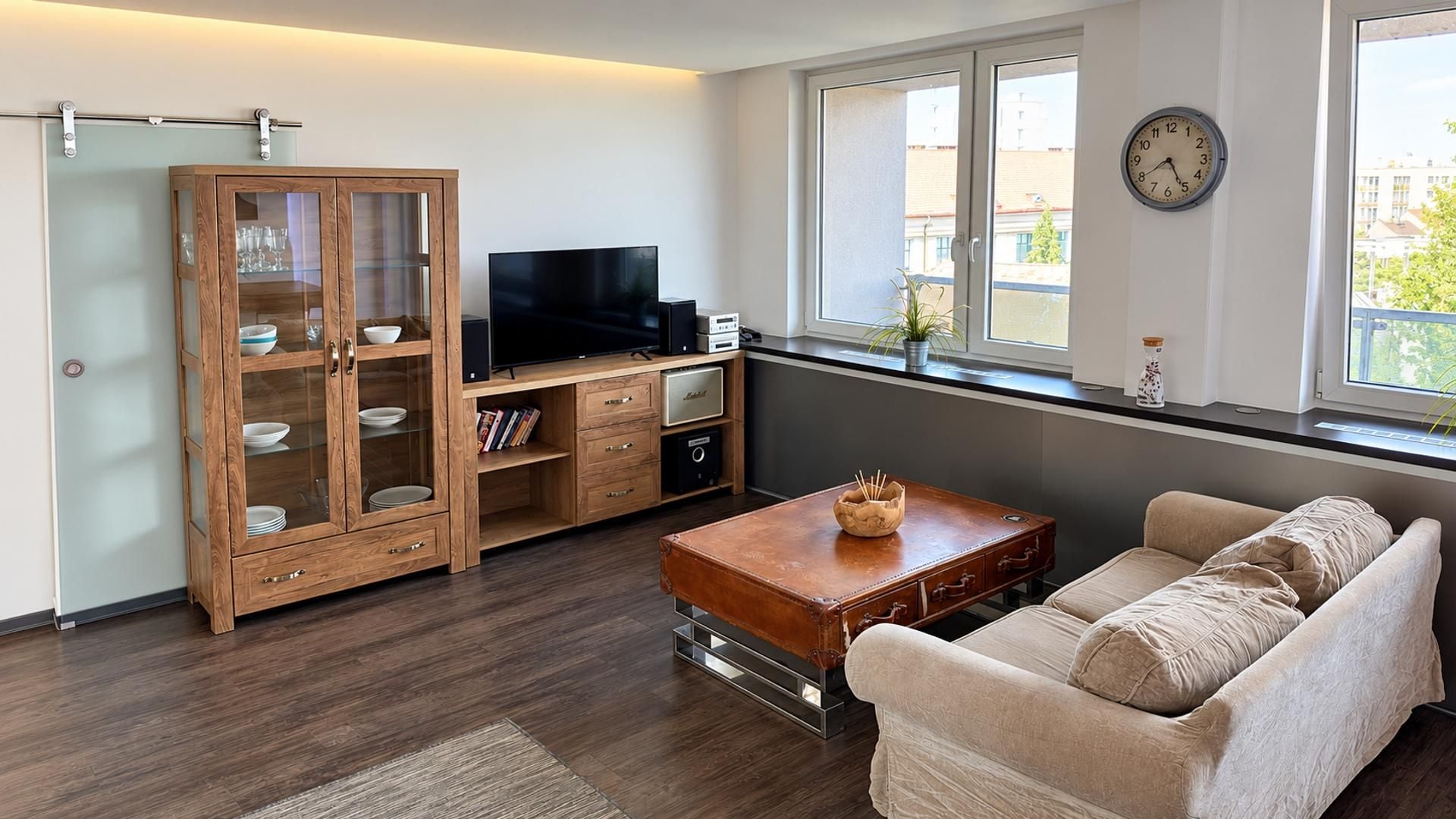 Ein Wohnzimmer mit einer Couch, einem Couchtisch, einem Fernseher, einer Vitrine und einem Fenster mit Blick auf Gebäude.