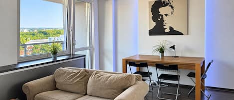 A modern living room with a sofa, a dining table with chairs, a potted plant, and a painting on the wall.