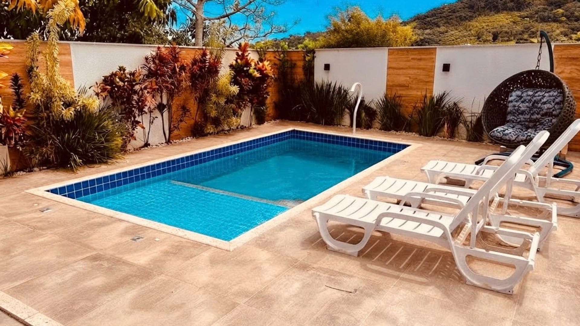 Ein rechteckiger Swimmingpool mit blauen Fliesen, umgeben von einer bepflasterten Liegefläche. Weiße Liegestühle und ein runder, gemusterter Stuhl sind vorhanden.