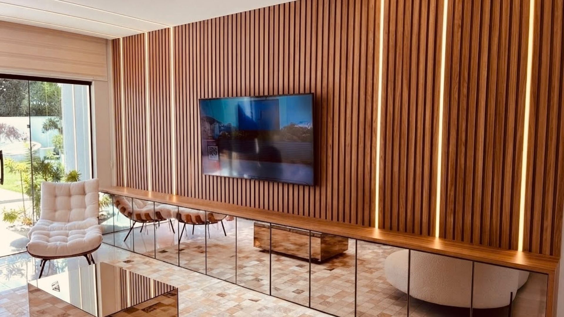 Una sala moderna con un televisor de pantalla plana montado en una pared de madera, una mesa de centro de vidrio y un sillón con cojines.