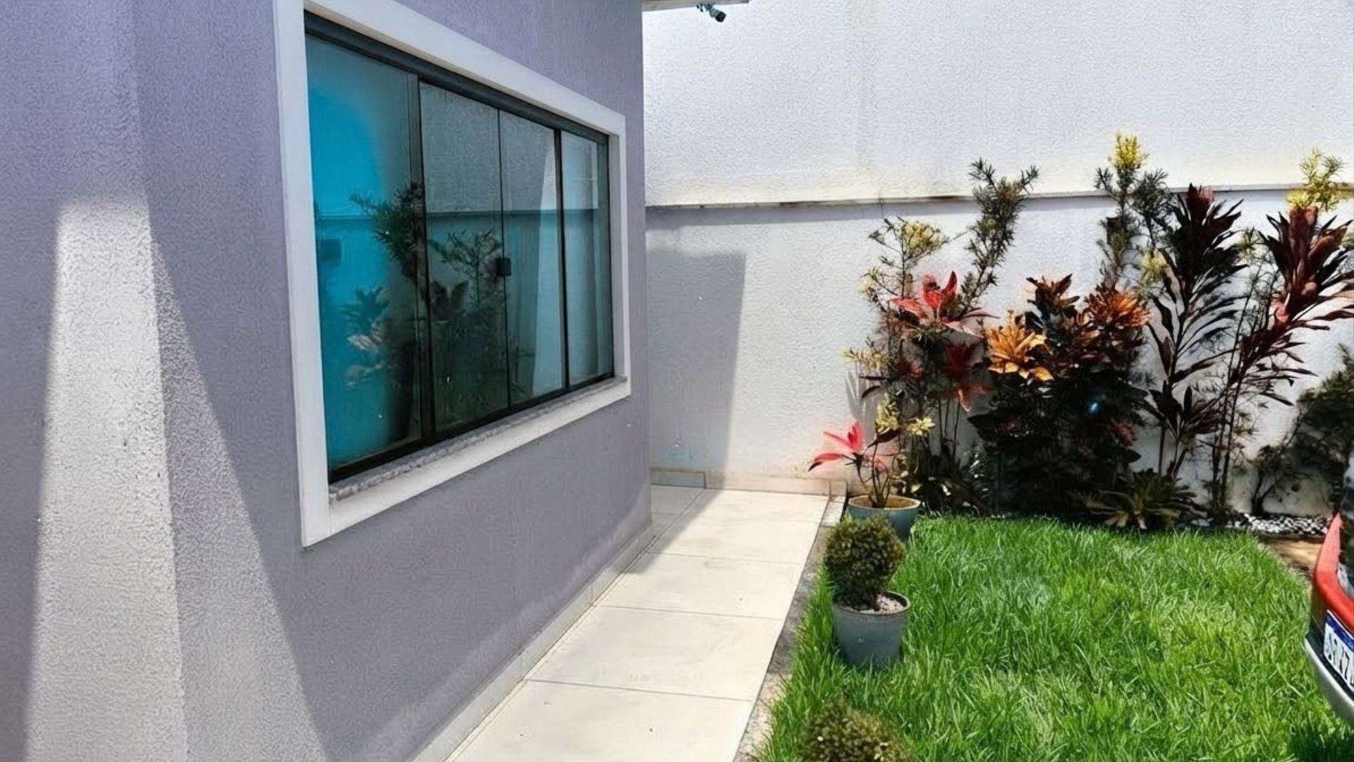 Un edificio moderno con un amplio ventanal, un pequeño jardín con diversas plantas y un cielo despejado.