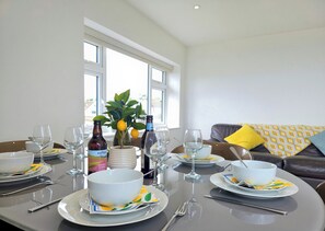 Ein Esstisch ist gedeckt mit Tellern, Schüsseln, Weingläsern und Besteck, im Hintergrund befinden sich ein Blumentopf und Flaschen.